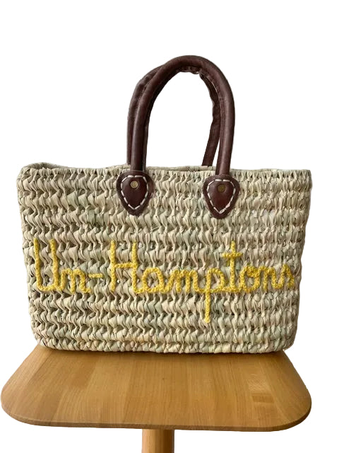 A woven straw tote bag with dark brown leather handles and yellow embroidered text reading 'Un-Hamptons' on the front, displayed on a wooden stool against a white embossed wall.